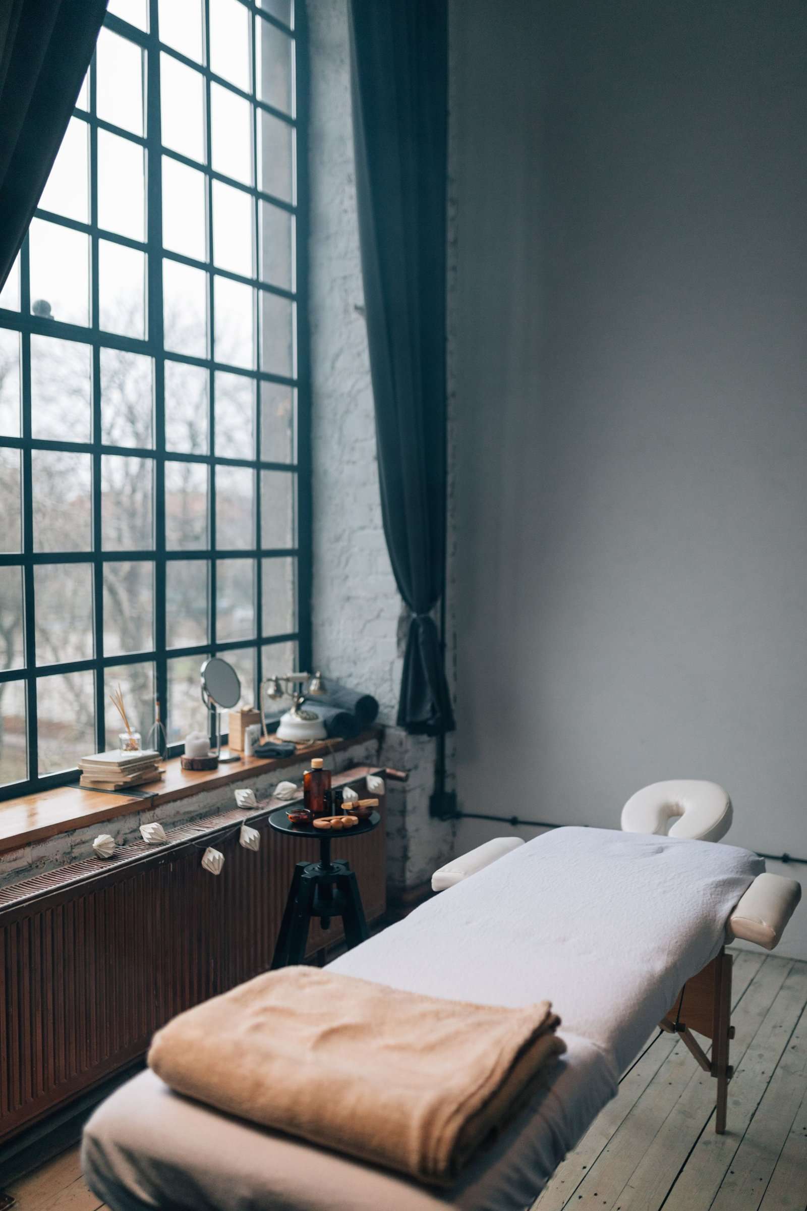 A bright treatment room with tall windows, a massage table and daylight