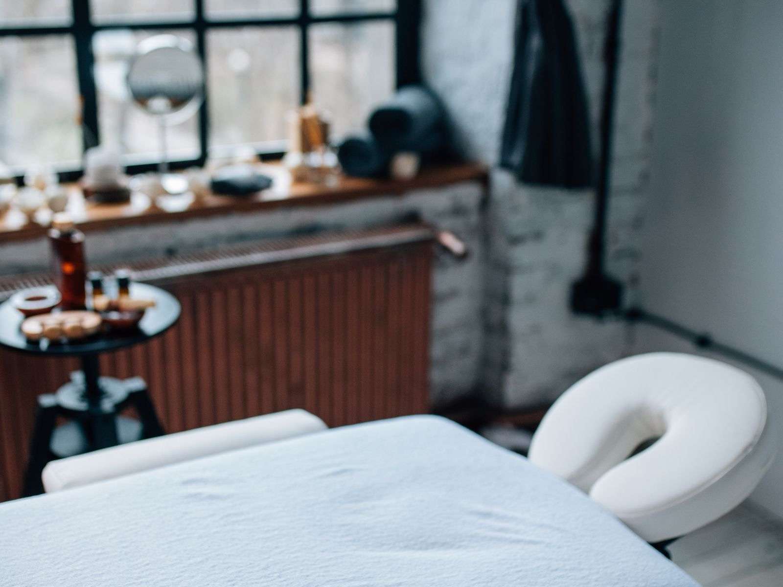 A bright treatment room with a massage table with a white sheet and a face cradle with an opening, in front of a window with a black frame